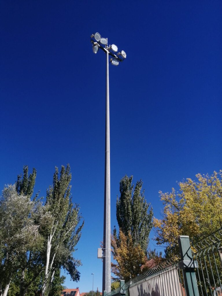 Farola alta con focos encendida en un entorno urbano con árboles y cielo azul despejado